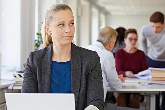 Yngre kvinna sitter vid en dator och tittar bort. I bakgrunden diskuterar kollegor. 
