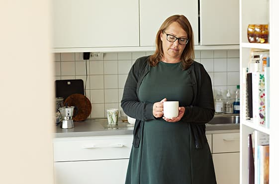 Kvinna står med kaffekopp i handen i ett kök, blicken nedslående