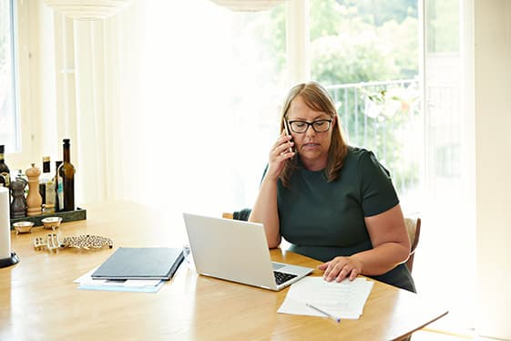 En kvinna sitter vid en dator och pratar i mobiltelefon. Hon är hemma vid ett köksbord.
